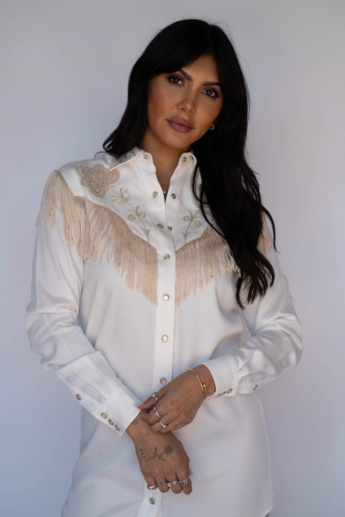 A woman with long dark hair wearing a white western-style button-up shirt with beige fringe and embroidery on the chest, posed against a plain light gray background.