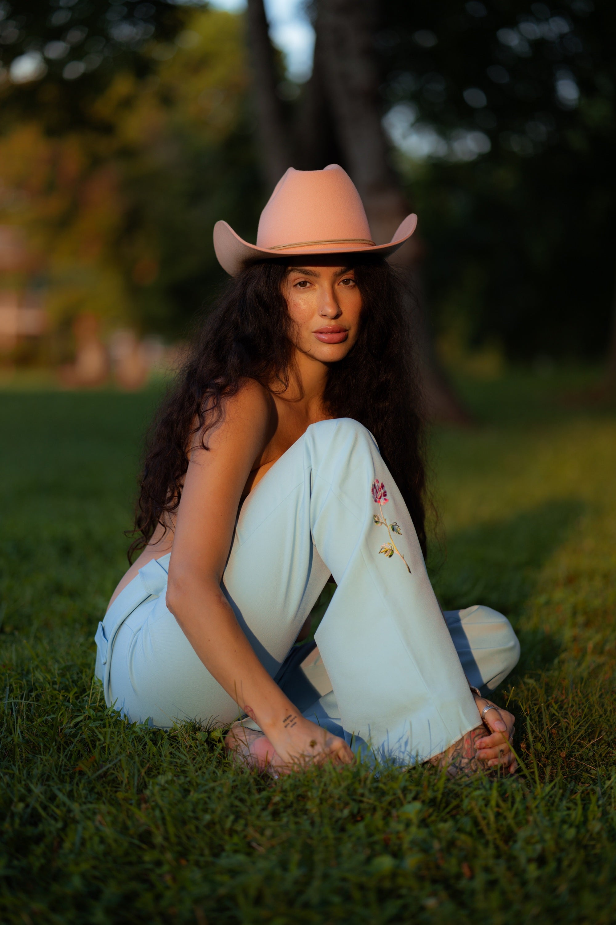 A woman sits cross-legged on green grass outdoors, wearing a light pink cowboy hat, pale blue wide-leg jeans with embroidered details, and sandals.