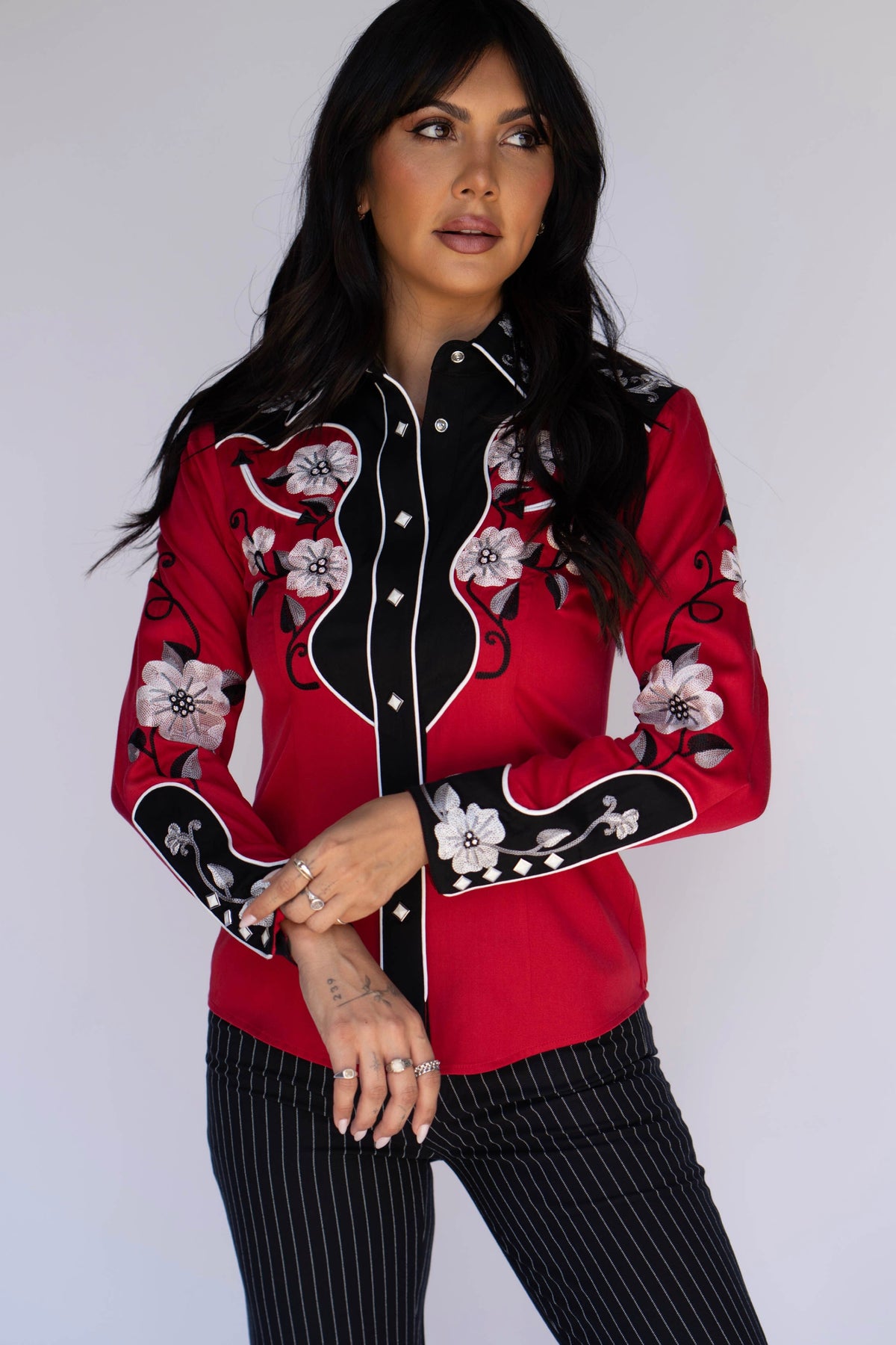 A woman modeling a red western-style button-up shirt with black and white floral embroidery, paired with black pinstriped pants, against a plain light gray background.