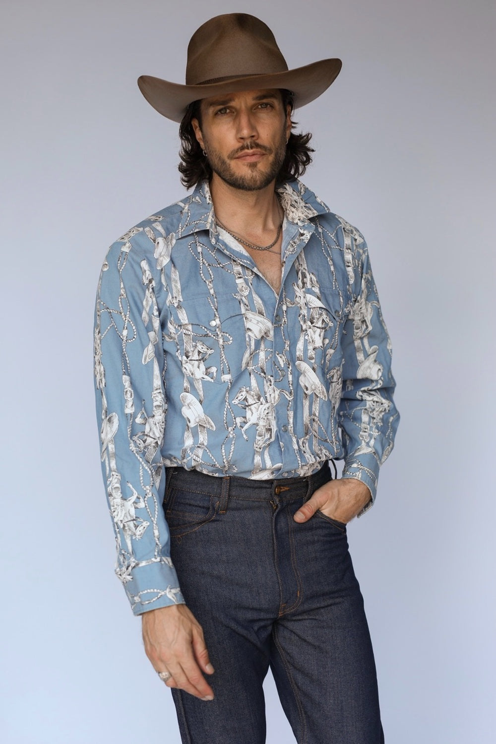A man wearing a light blue western-style button-down shirt with a white and gray bird print, dark blue jeans, and a brown cowboy hat, standing against a light gray studio background.