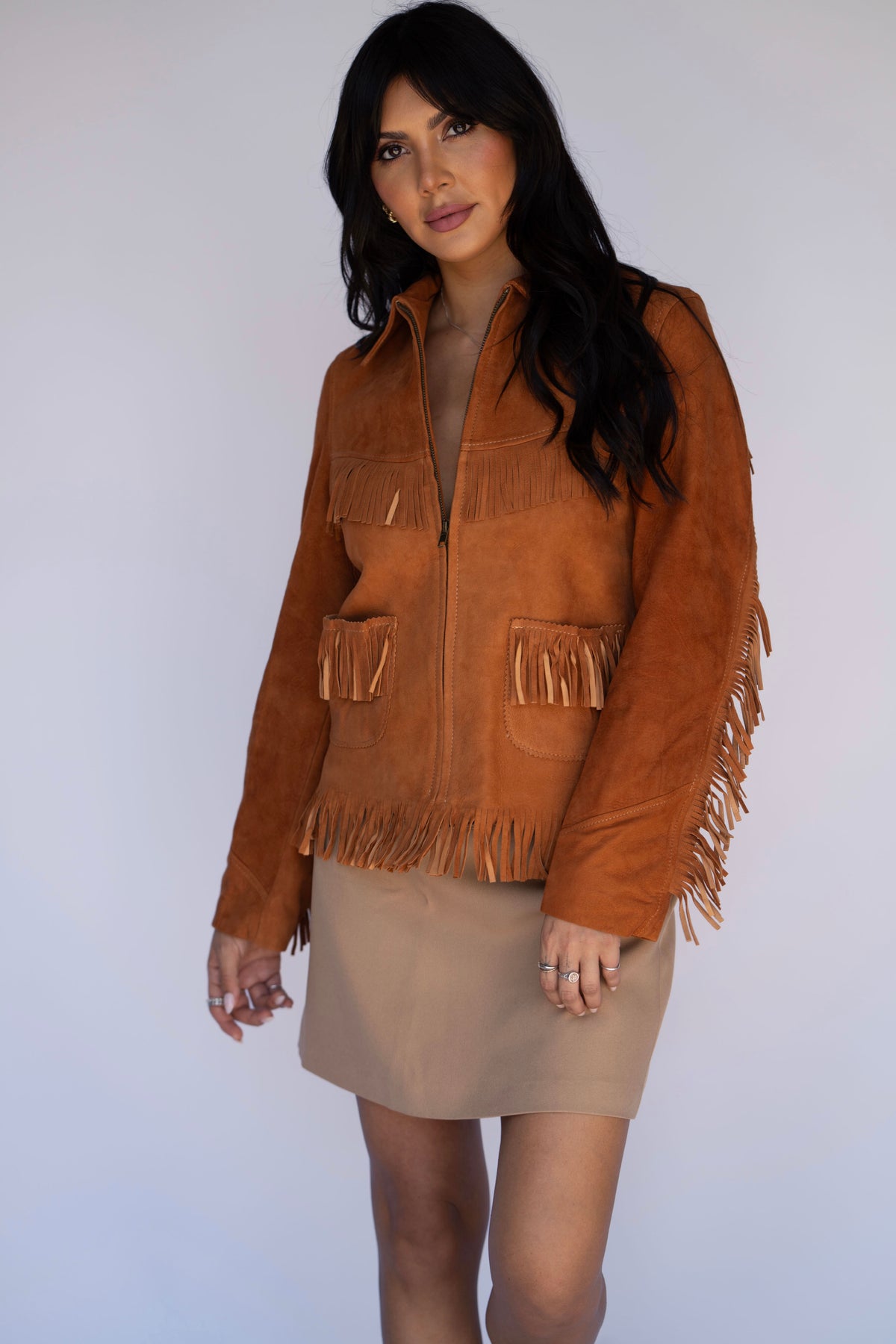 A woman modeling a brown suede fringe jacket with a front zipper and pockets, paired with a beige mini skirt, against a plain light gray studio background.