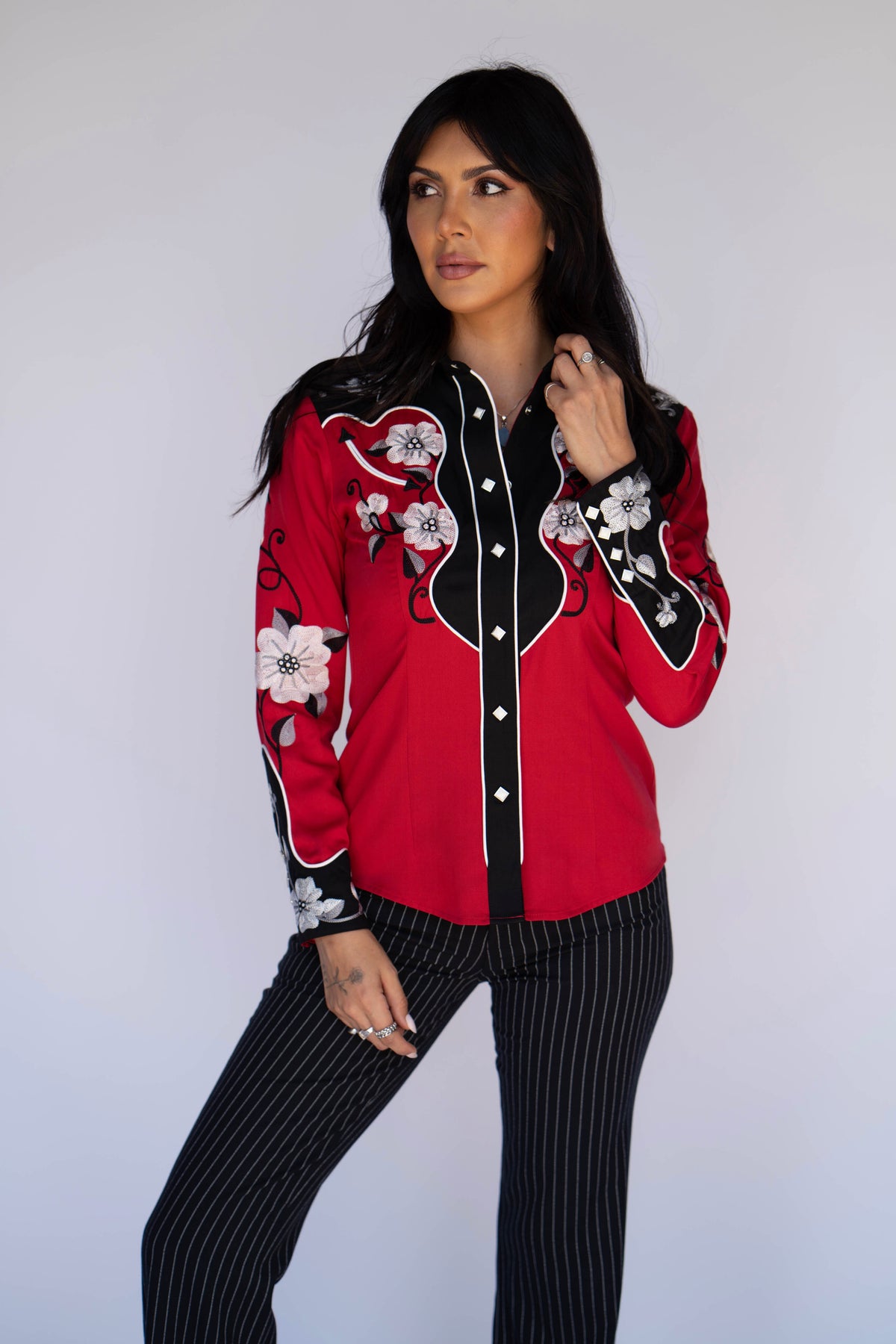 A woman modeling a red western-style button-up shirt with black yoke panels, white floral embroidery, and black-and-white pinstriped pants against a plain light gray background.