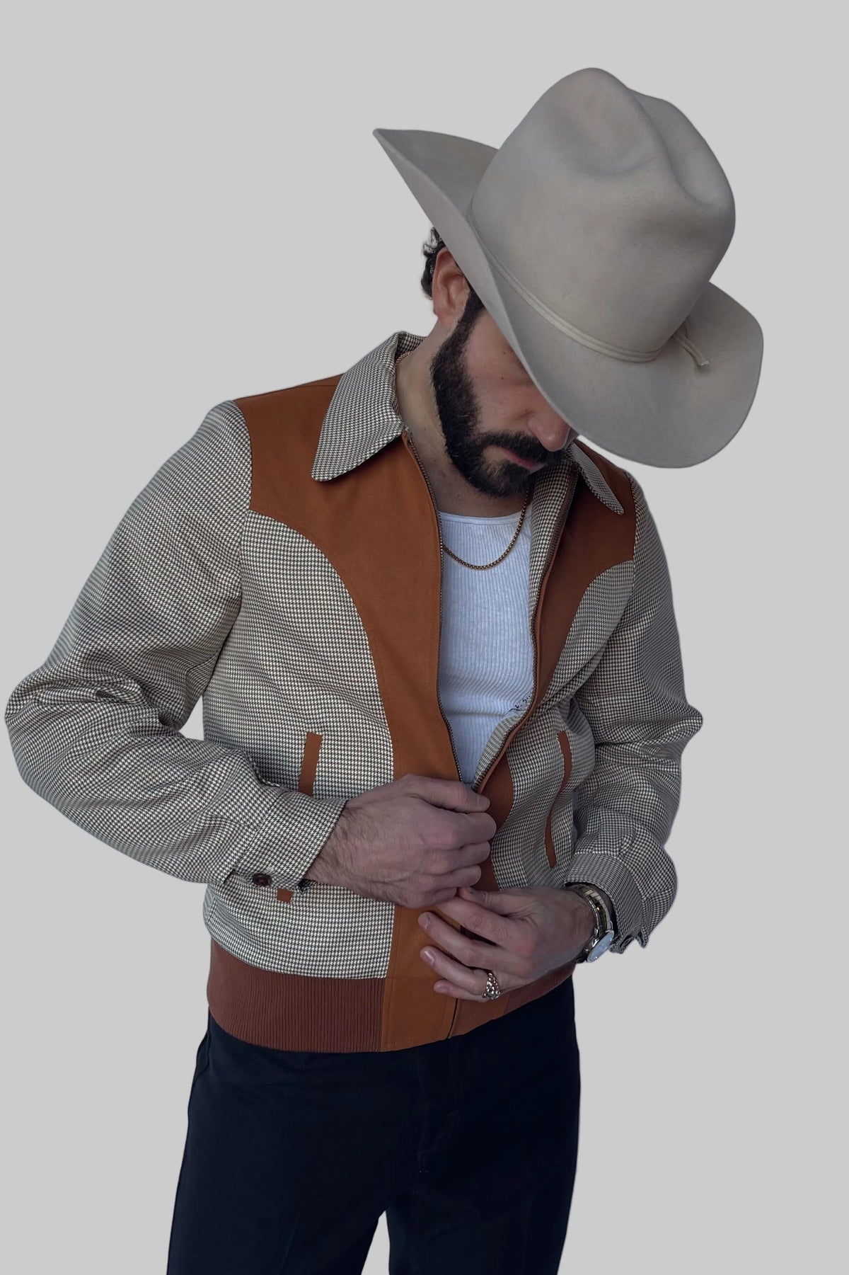 A man wearing a beige cowboy hat, a beige and brown color-blocked jacket over a white t-shirt, and dark pants, posing against a solid gray background.