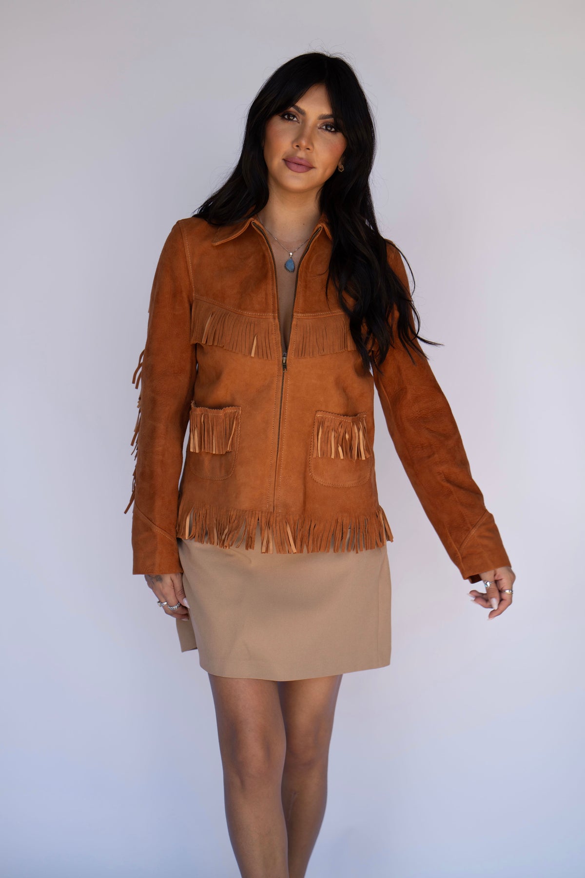 A woman modeling a brown suede fringe jacket over a beige mini skirt, standing against a plain light gray studio background.