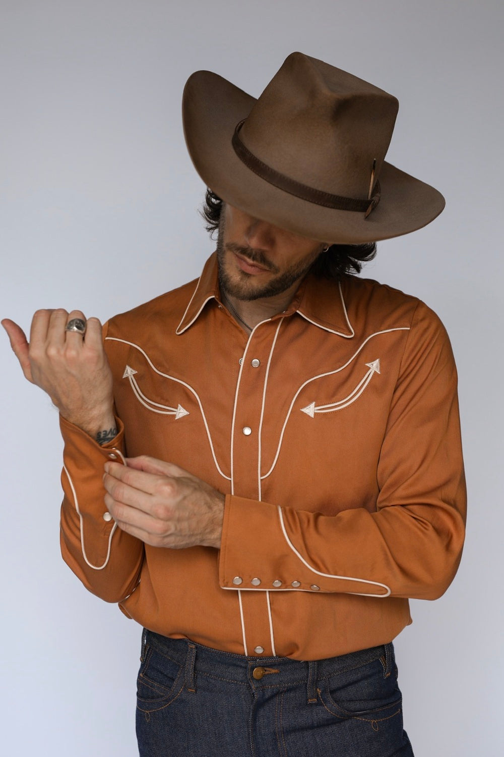 A man wearing a brown cowboy hat and a rust-orange western long-sleeve shirt with white contrast stitching, paired with dark blue jeans, photographed against a light gray studio background.