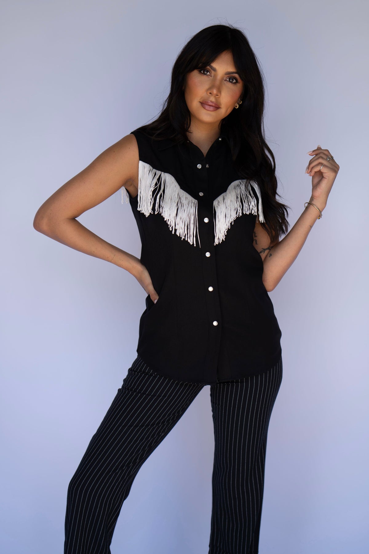 A woman modeling a black sleeveless western-style button-up shirt with white fringe across the chest, paired with black pinstriped pants, against a light blue studio background.