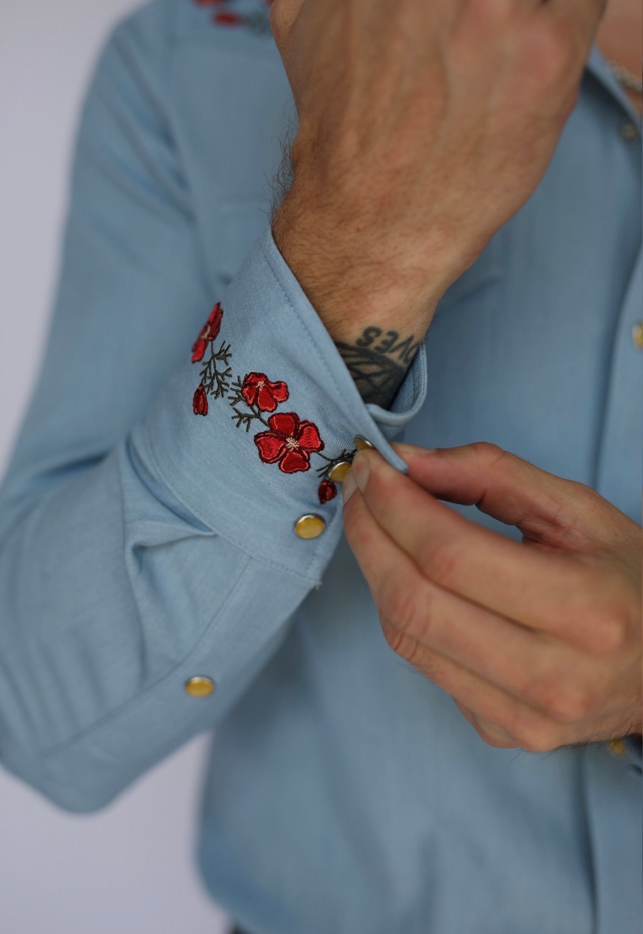 Close-up of a person wearing a light blue dress shirt with red floral embroidery on the cuff and gold buttons, adjusting the cuff with one hand.