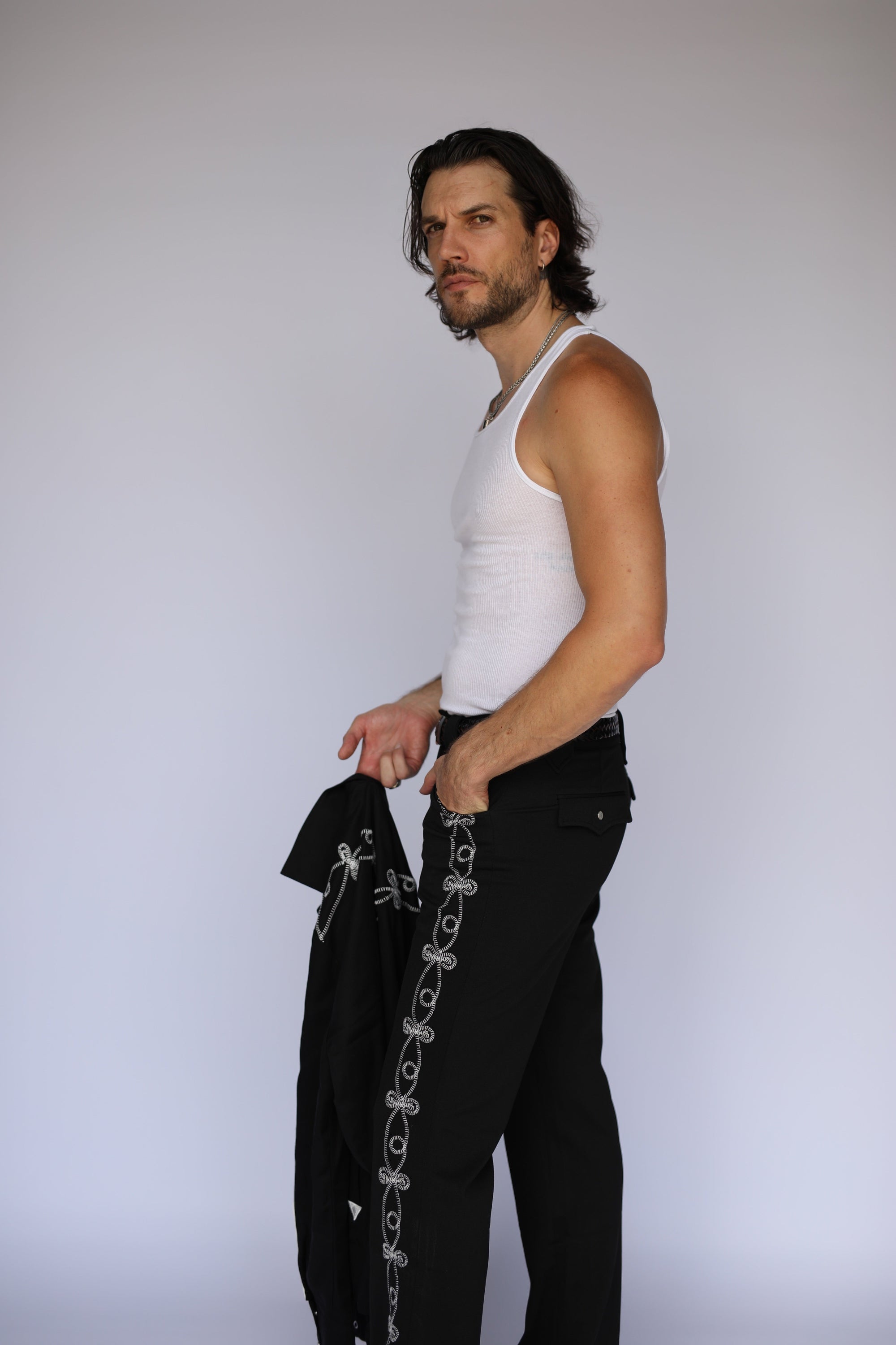 A man in a white tank top and black pants with silver chain detailing along the side, holding a black jacket with silver chain details, standing against a light gray studio background.