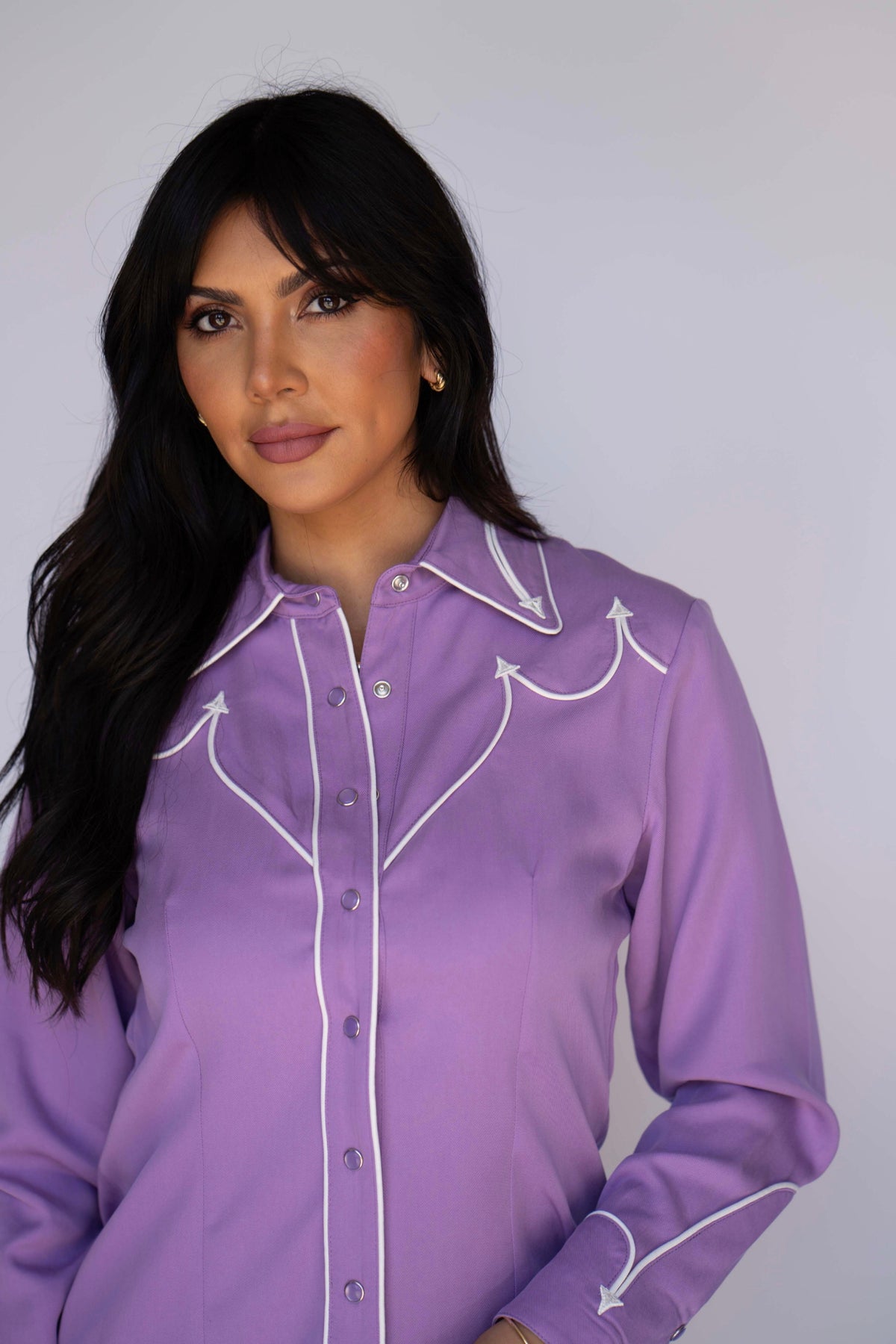 A woman wearing a lavender purple long-sleeve western-style button-up shirt with white embroidered details on the chest and cuffs, photographed against a plain light gray background.