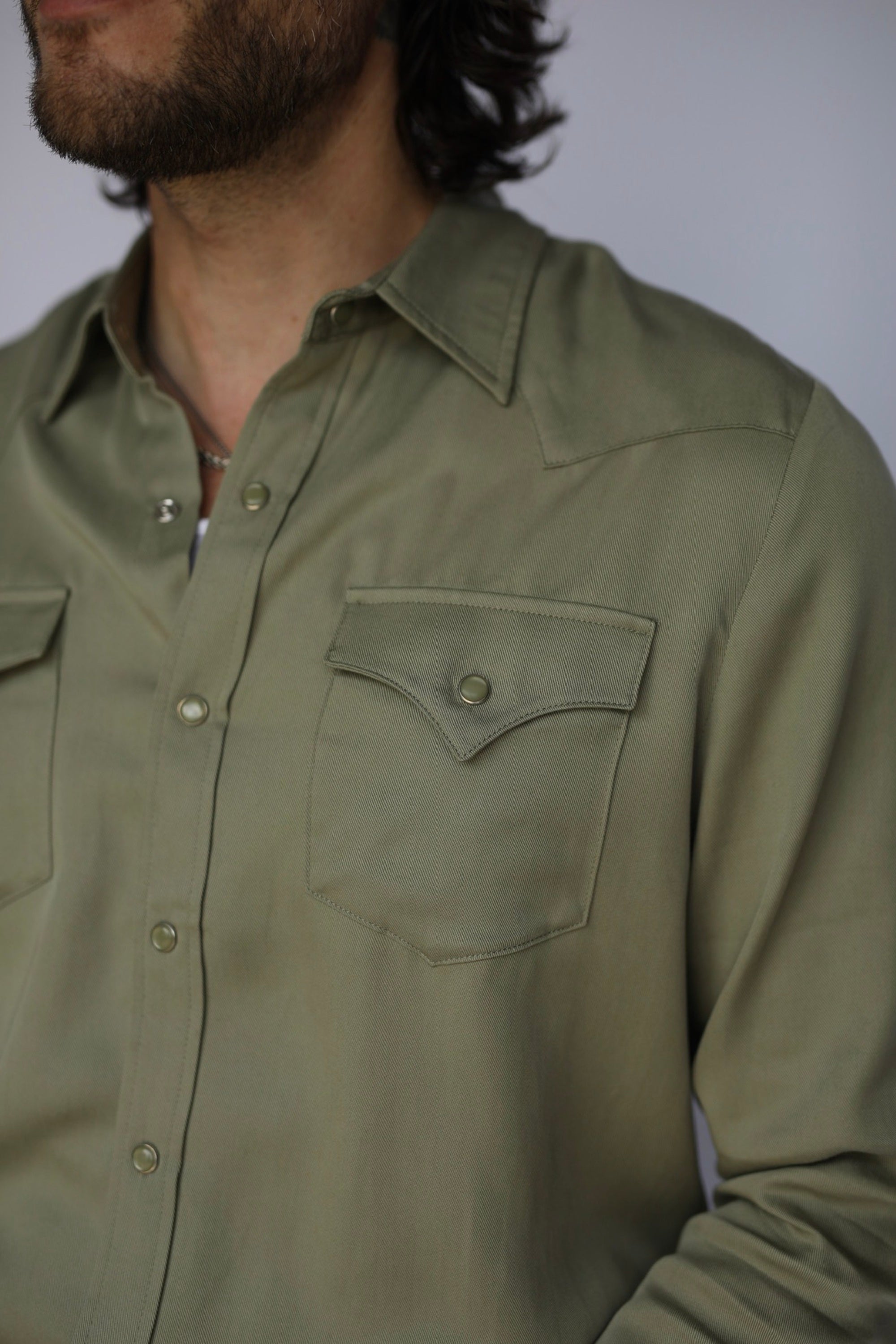 Close-up of a man wearing a muted olive green western-style button-down shirt with chest pockets, showing the collar, buttons, and fabric texture.
