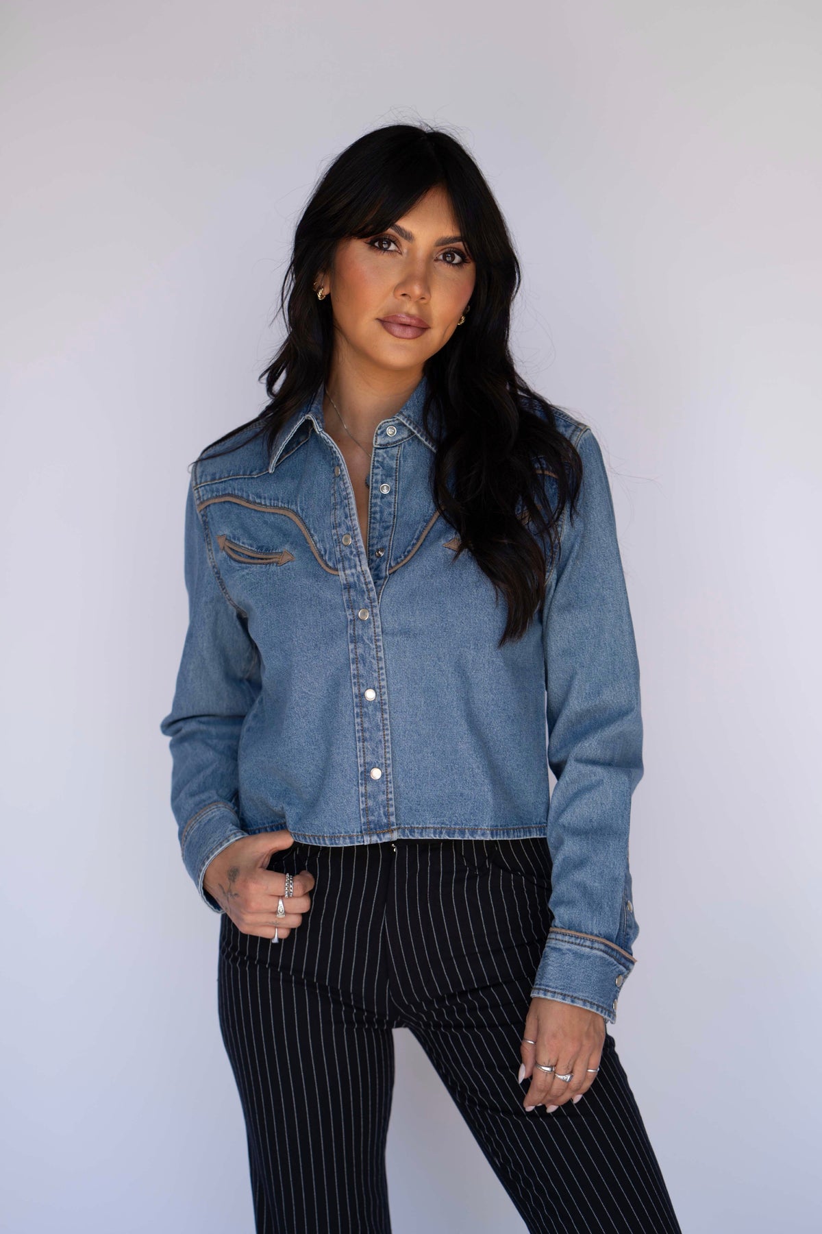 A woman modeling a light blue cropped denim shirt-jacket with brown contrast stitching, paired with black pinstriped pants, against a plain light gray background.