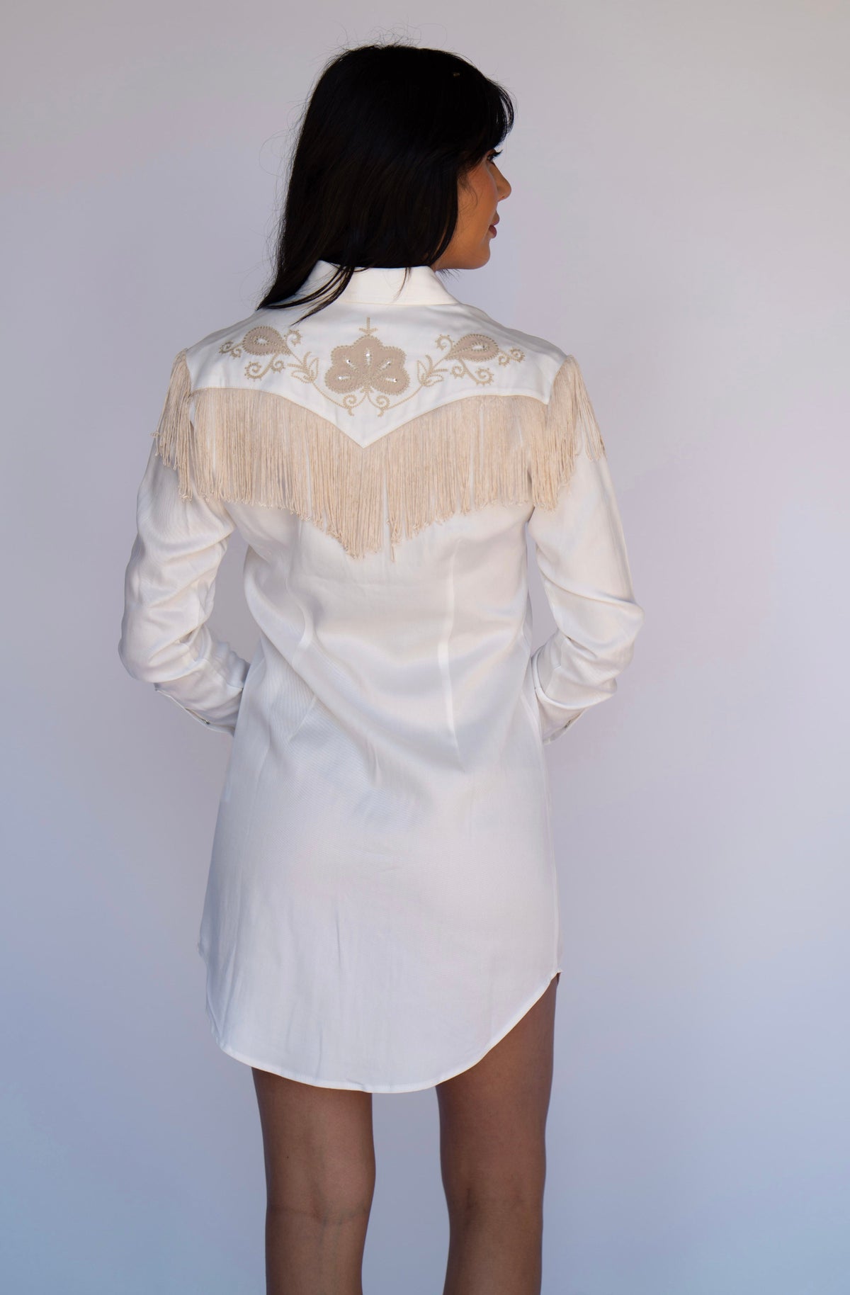 A woman shown from the back wearing a white long-sleeve western-style shirt dress with beige fringe and embroidery across the shoulders, against a plain light gray background.