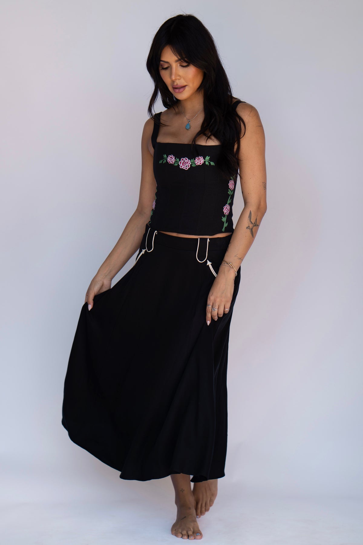 A woman modeling a black two-piece outfit consisting of a sleeveless crop top with pink and green floral embroidery and a matching long black skirt with a drawstring waist, set against a plain light background.