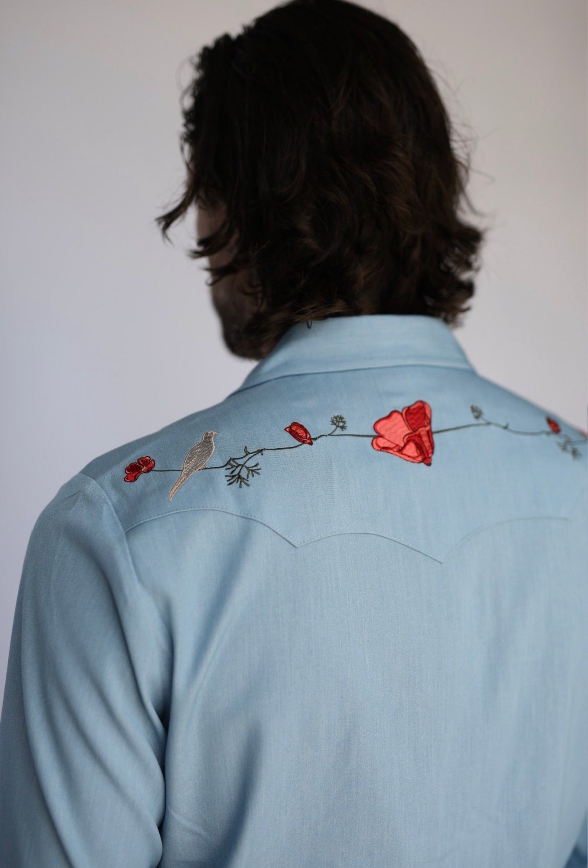 A man viewed from behind wearing a light blue western-style shirt with embroidered floral and bird details across the upper back.