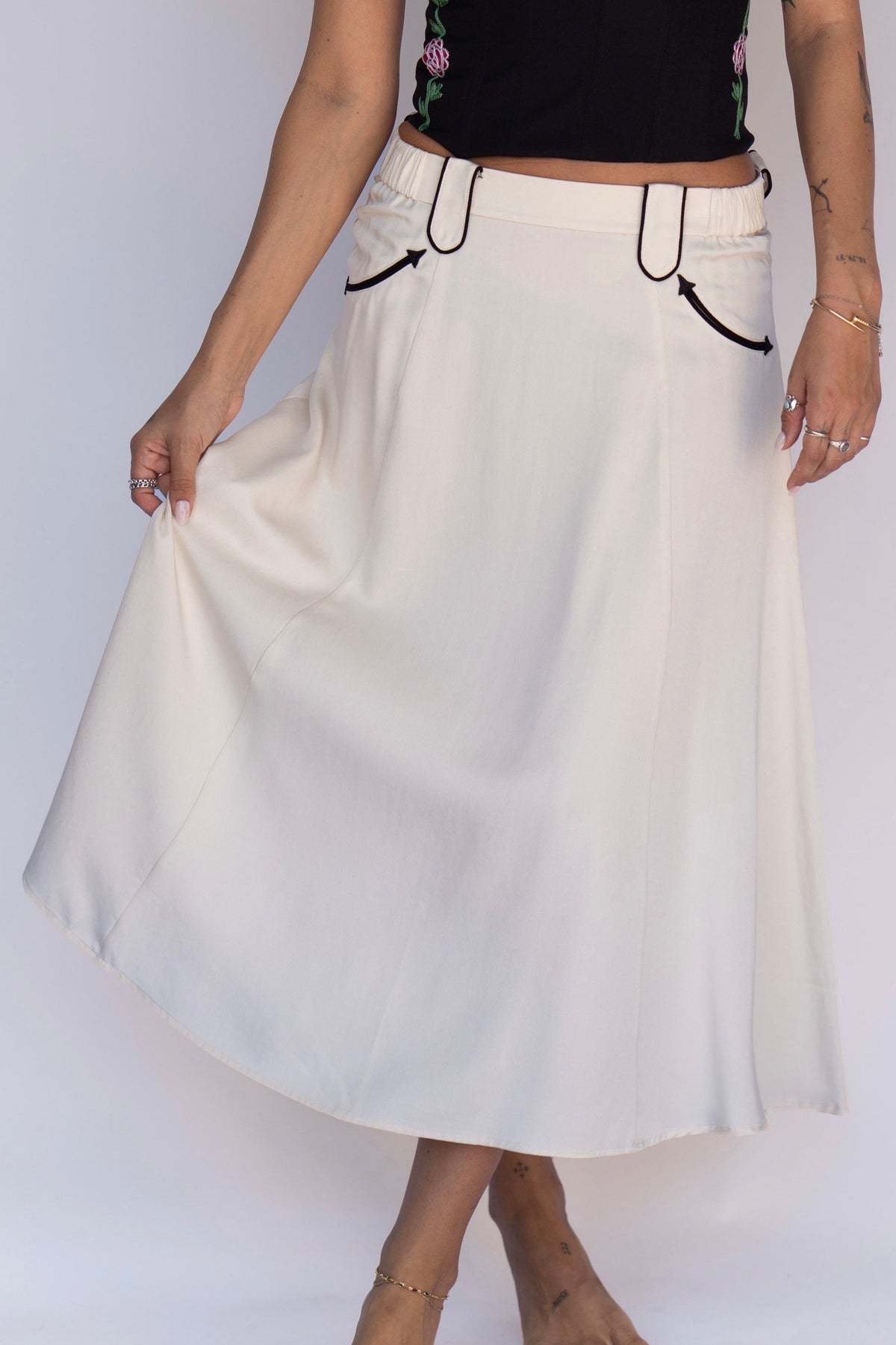 A woman modeling a black sleeveless crop top with pink floral embroidery and a long cream-colored skirt, standing against a plain white studio background.