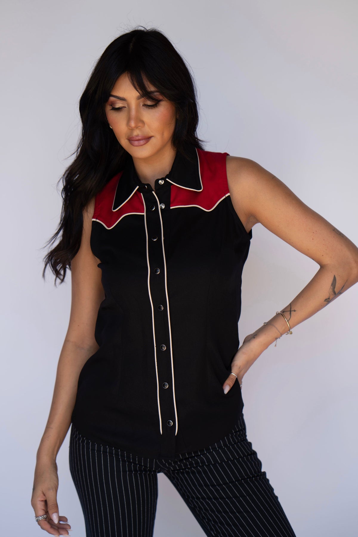A woman modeling a sleeveless black button-front top with a red and black western-style collar and white piping, paired with black pinstriped pants, against a plain light gray background.