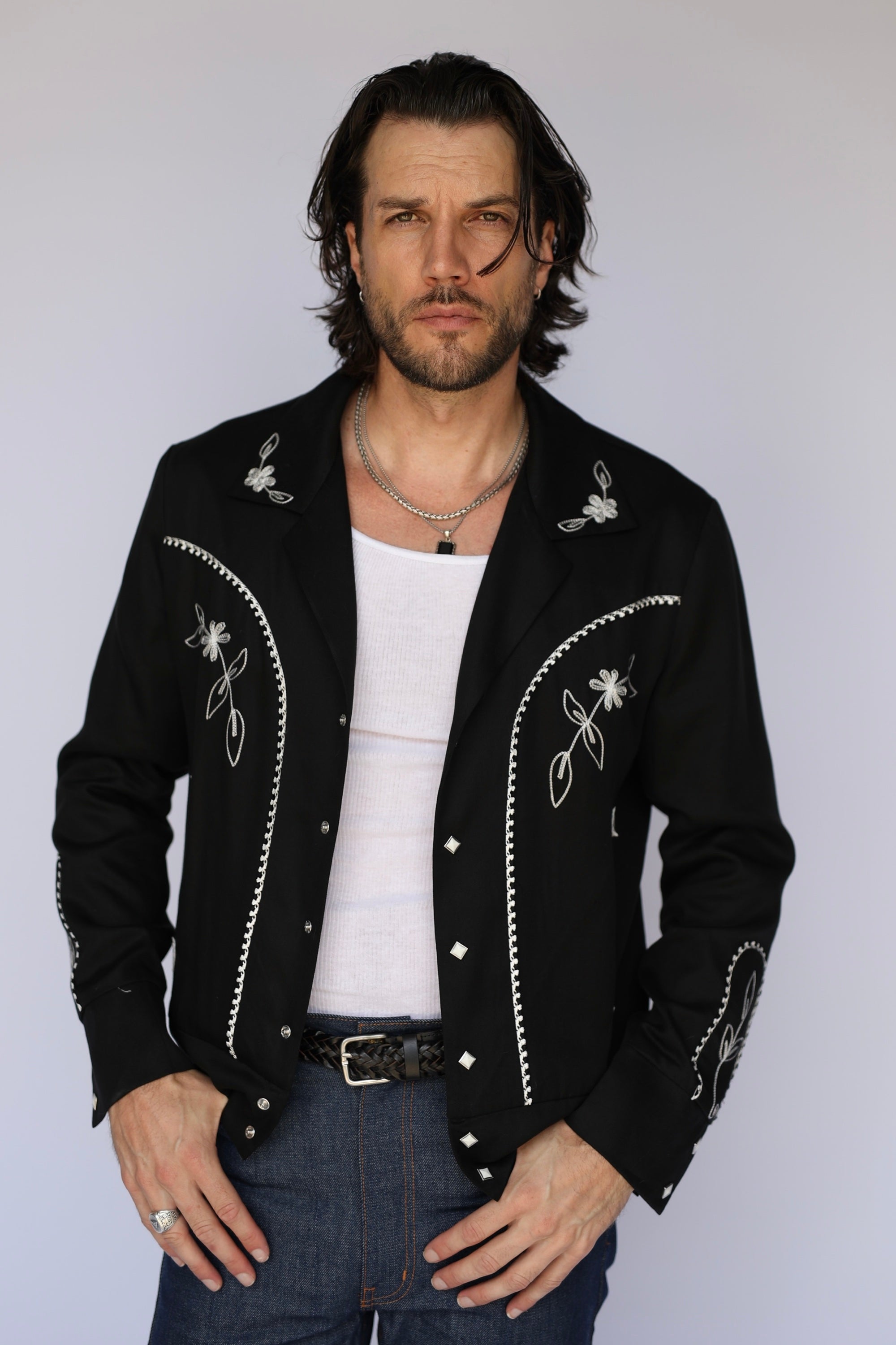 A man wearing a black western-style jacket with white embroidered details and silver studs, layered over a white tank top and blue jeans, photographed against a light gray studio background.