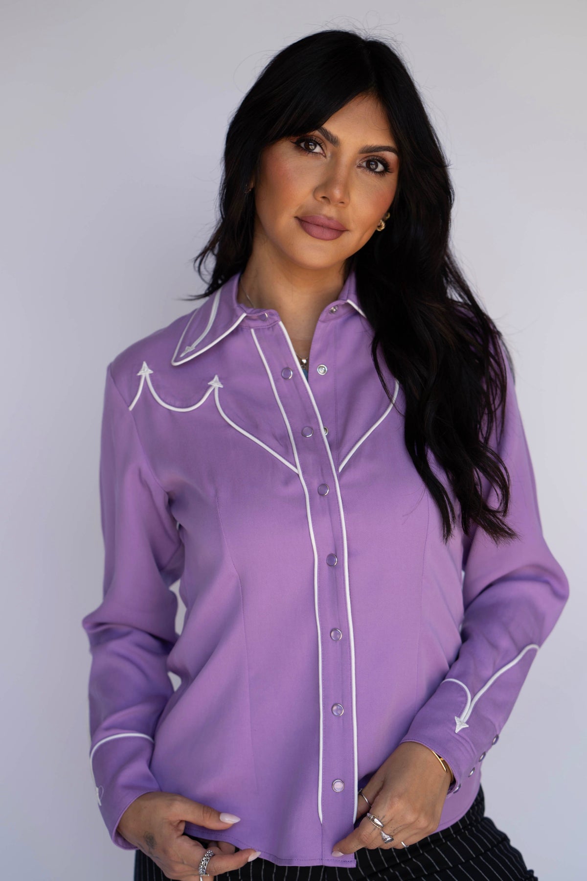 A woman modeling a lavender purple western-style long-sleeve button-up shirt with white embroidered details and piping, set against a plain light gray background.