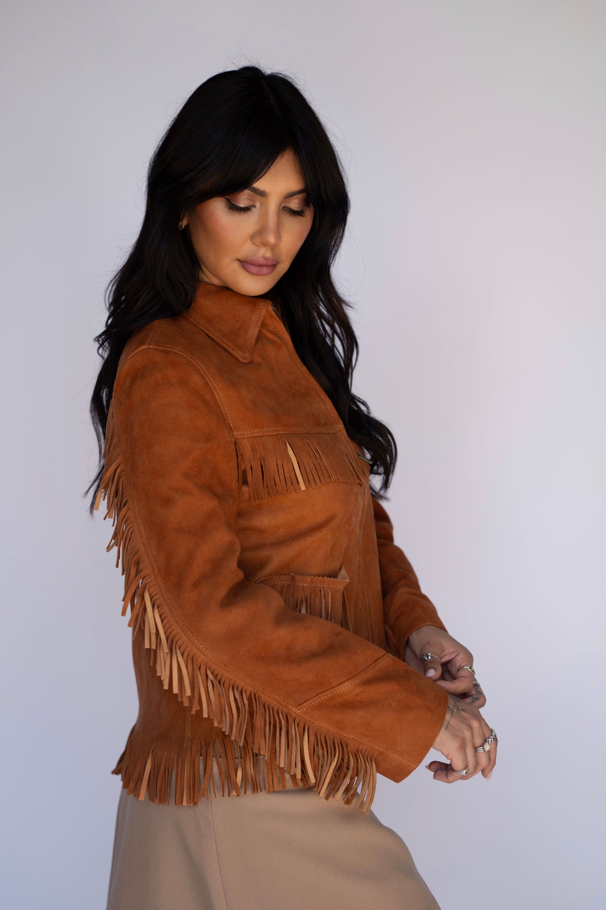 A woman wearing a brown suede fringe jacket and a beige skirt, posing against a light gray studio background.