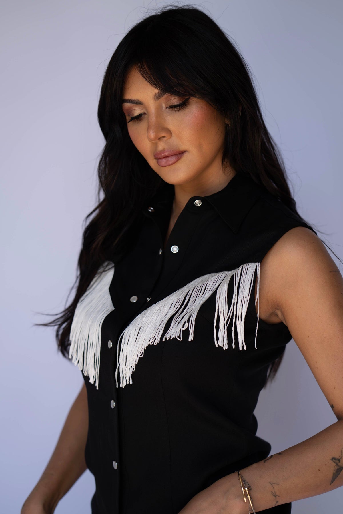 A woman wearing a sleeveless black button-front top with white fringe across the chest, photographed against a plain light background.