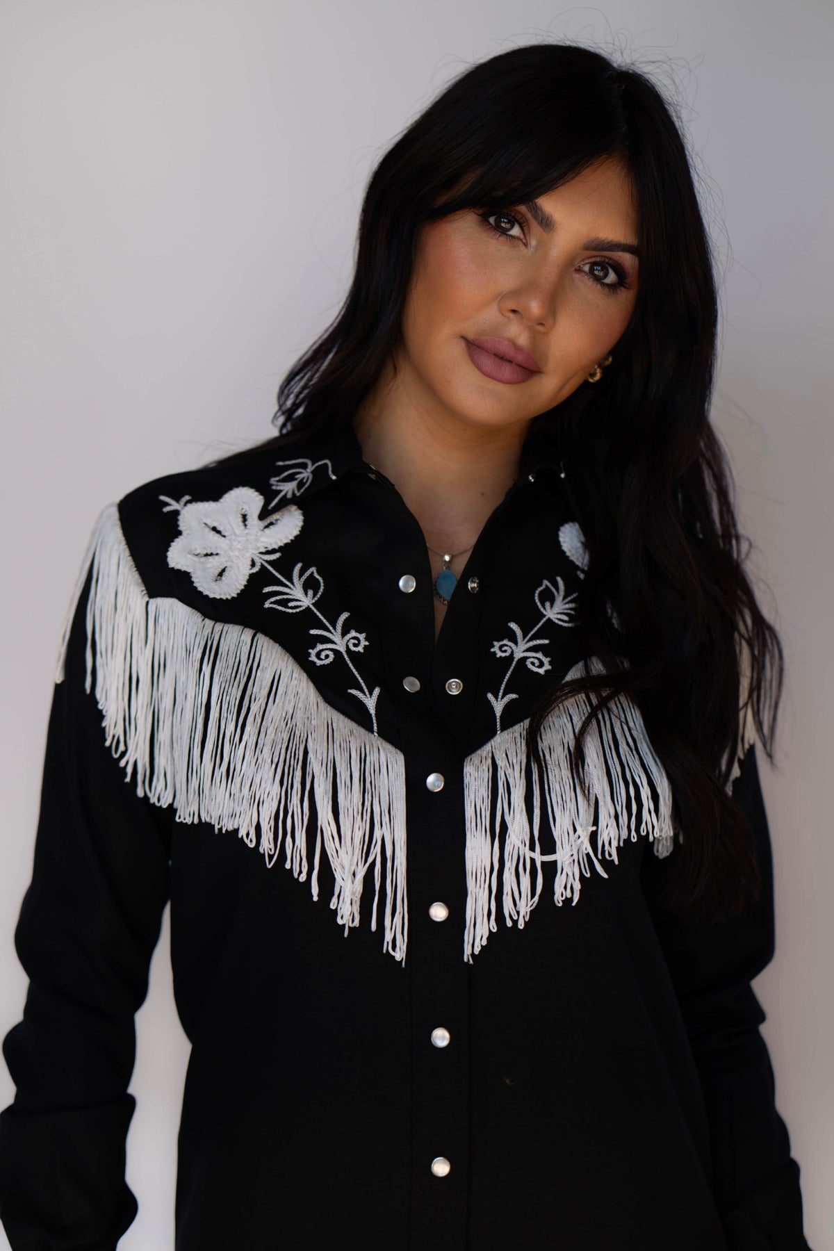 A woman wearing a black western-style button-up shirt with white floral embroidery and large white fringe across the chest, photographed against a plain light gray background.