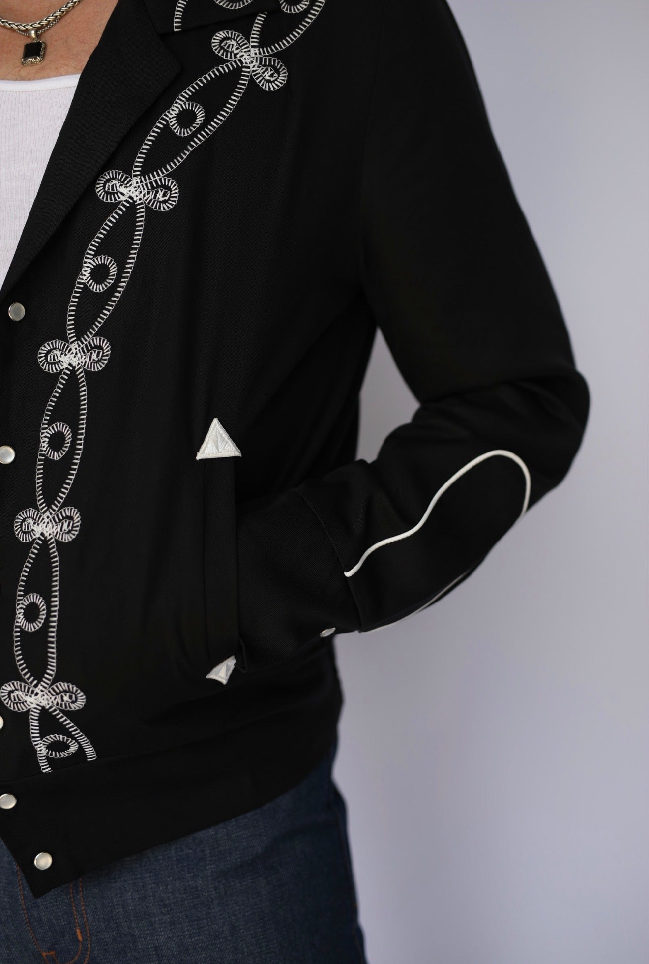 A close-up of a black western-style jacket with intricate silver rhinestone embroidery along the front and sleeve, worn by a model with blue jeans.