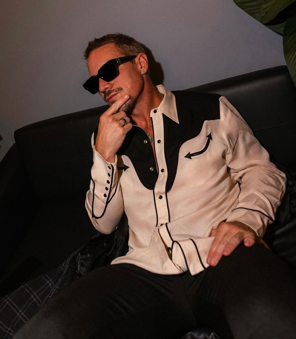 Diplo is wearing a stylish outfit with a white shirt and black accents, sitting on a dark couch.