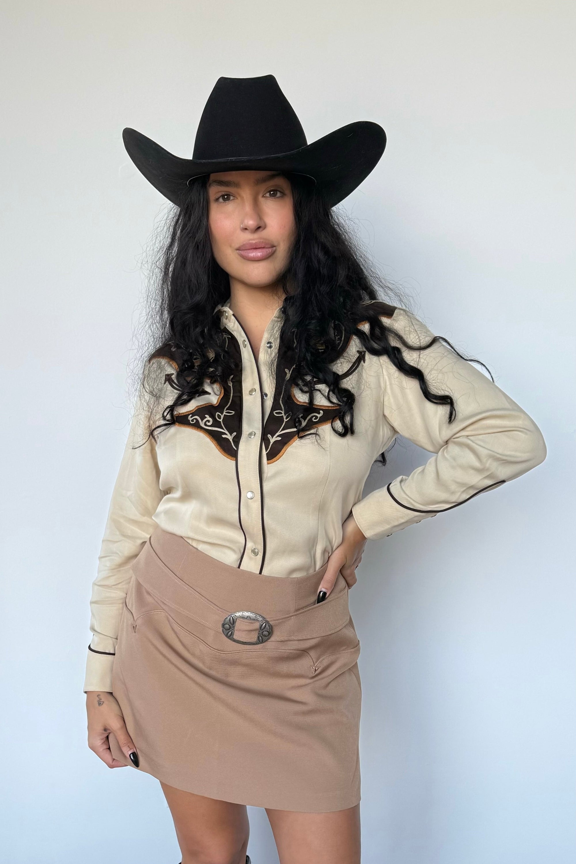Woman wearing a beige long-sleeve shirt, tan skirt, and black cowboy hat against a white background