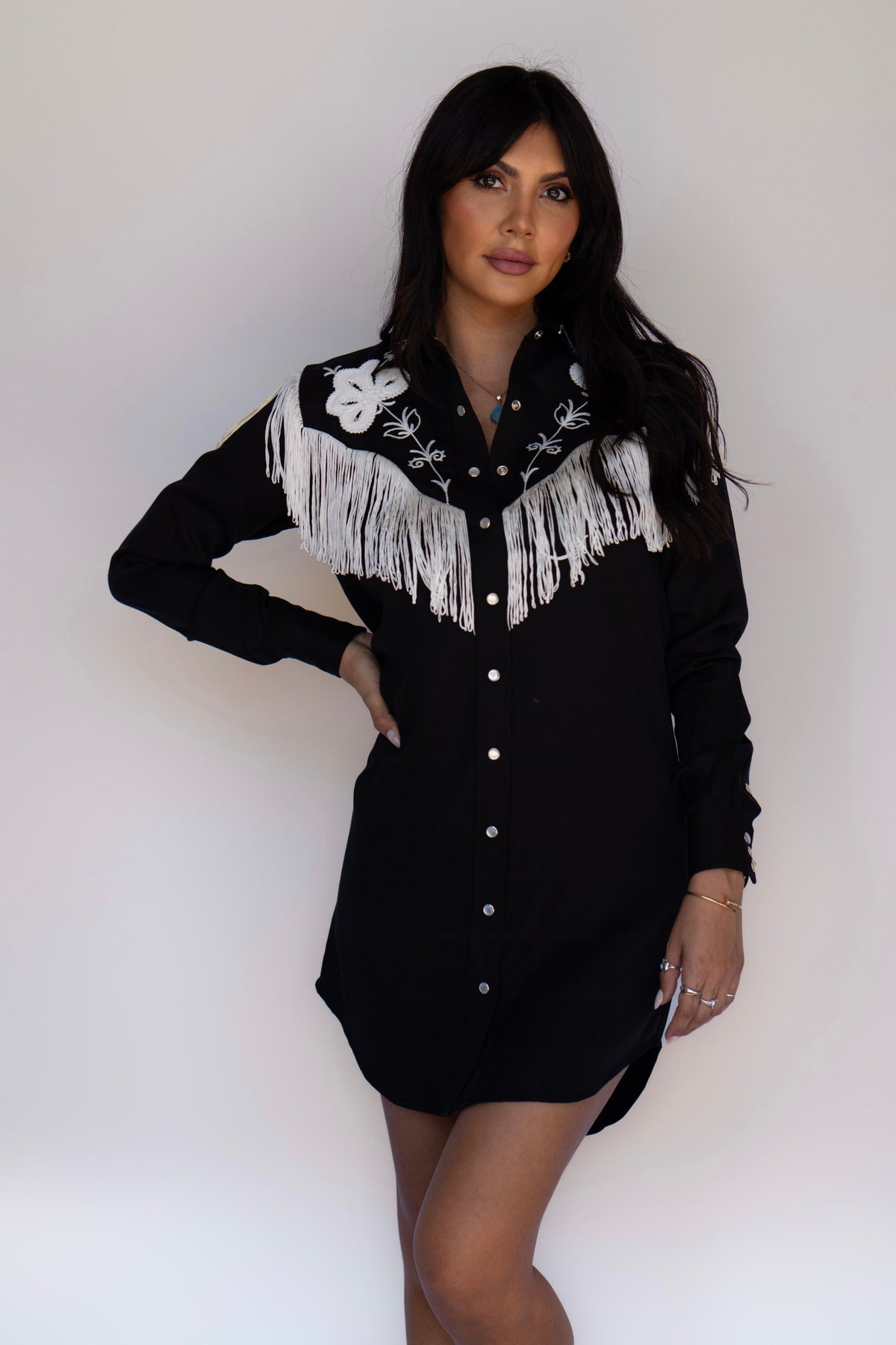 A woman modeling a black western-style shirt dress with white fringe and embroidery on the yoke, posed against a plain white studio background.