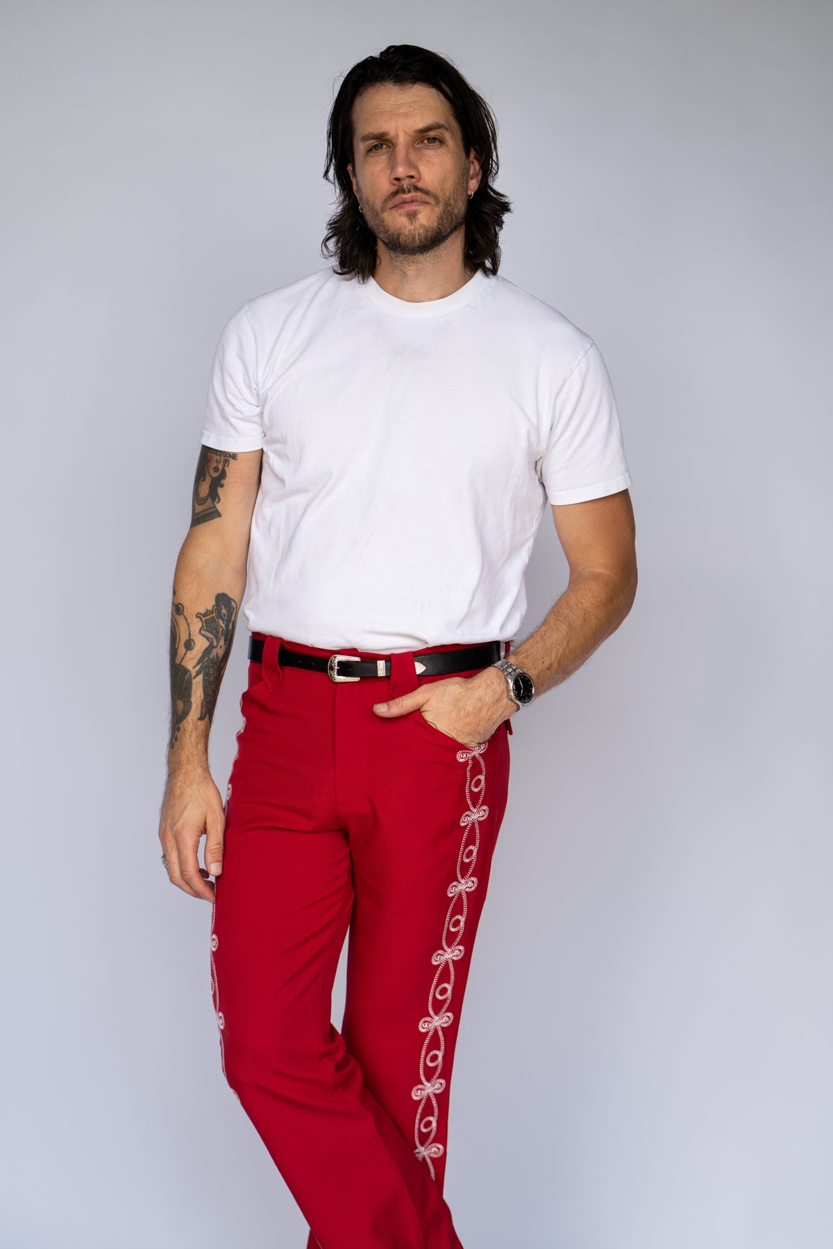A man modeling bright red pants with white decorative embroidery down the side, paired with a white t-shirt and a black belt, against a light gray background.
