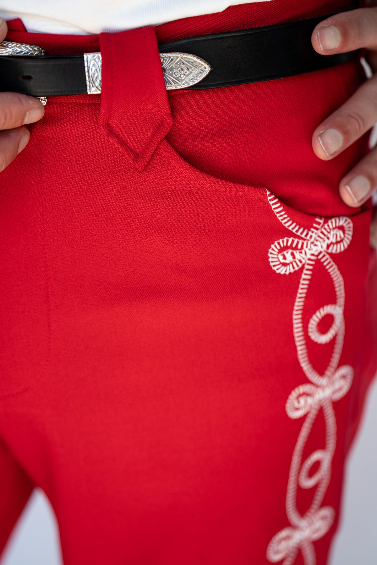 Close-up of bright red western-style pants with white embroidered scrollwork down the side, worn with a black belt featuring a silver buckle.
