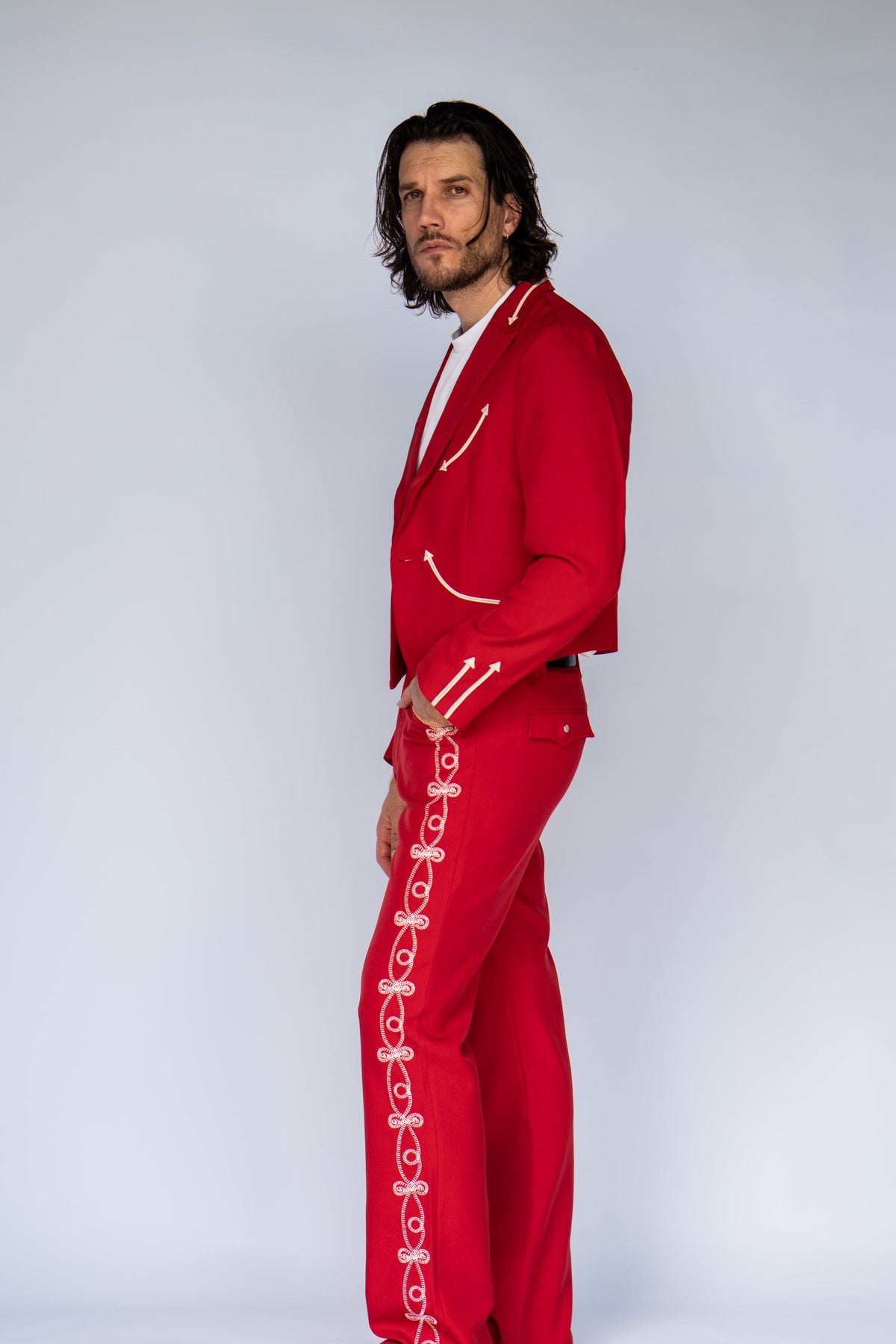 A man modeling a bright red two-piece tracksuit with white decorative trim and white rope details down the front of the pants, posed against a light gray studio background.