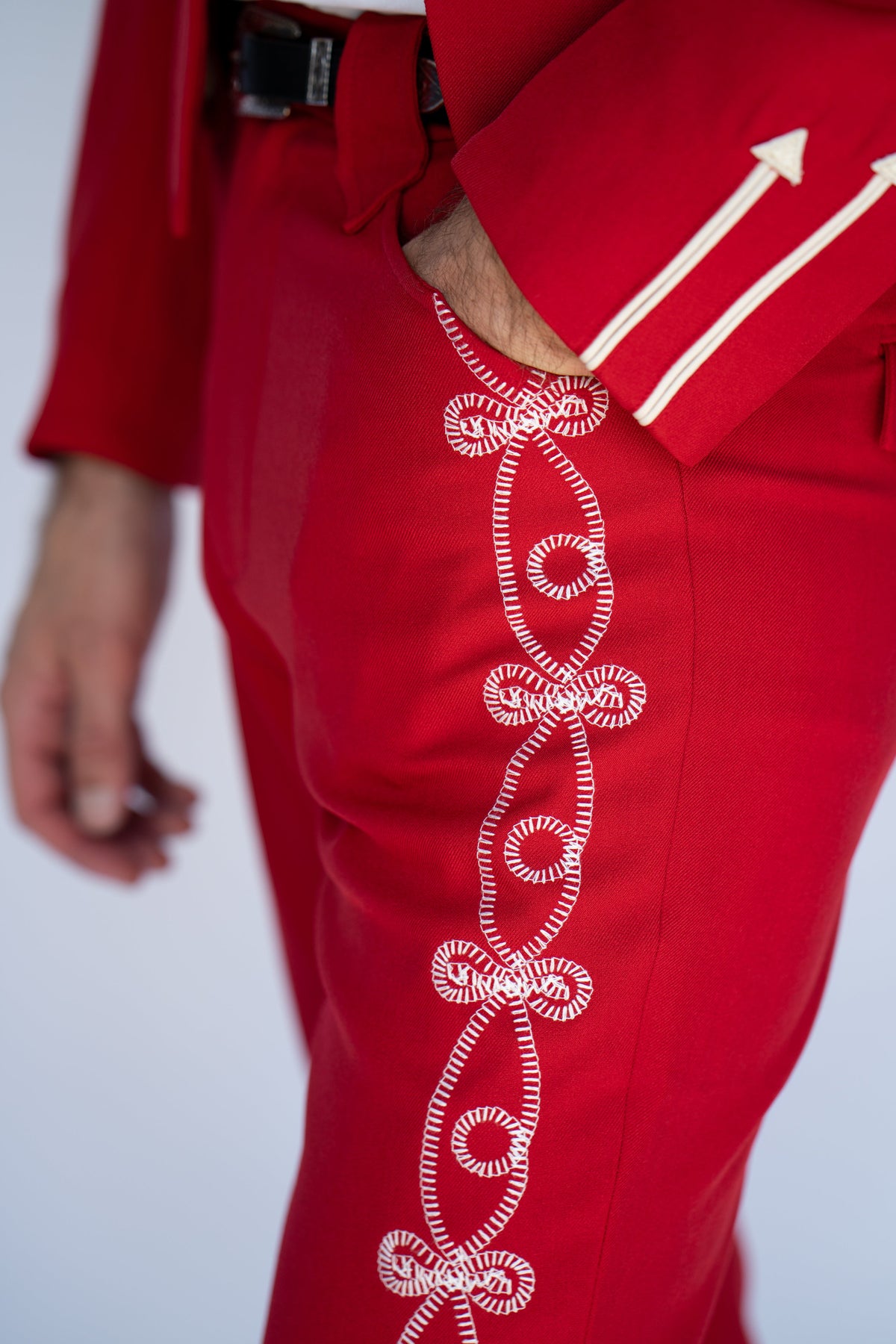 Close-up of a person wearing bright red tailored trousers with white decorative embroidered detailing running down the side, paired with a matching red jacket or shirt. The hand in the pocket is visible, and the background is a clean light gray studio backdrop.