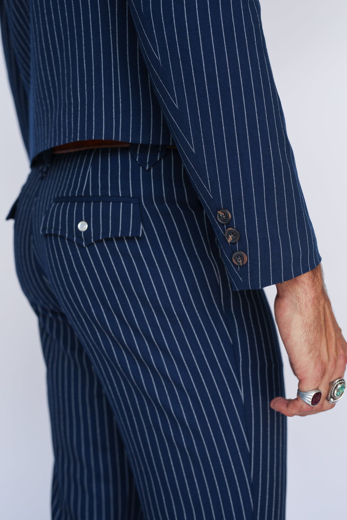 Close-up of a person wearing a navy blue pinstripe suit, showing the back pocket, waist, and hand with rings against a light gray studio background.