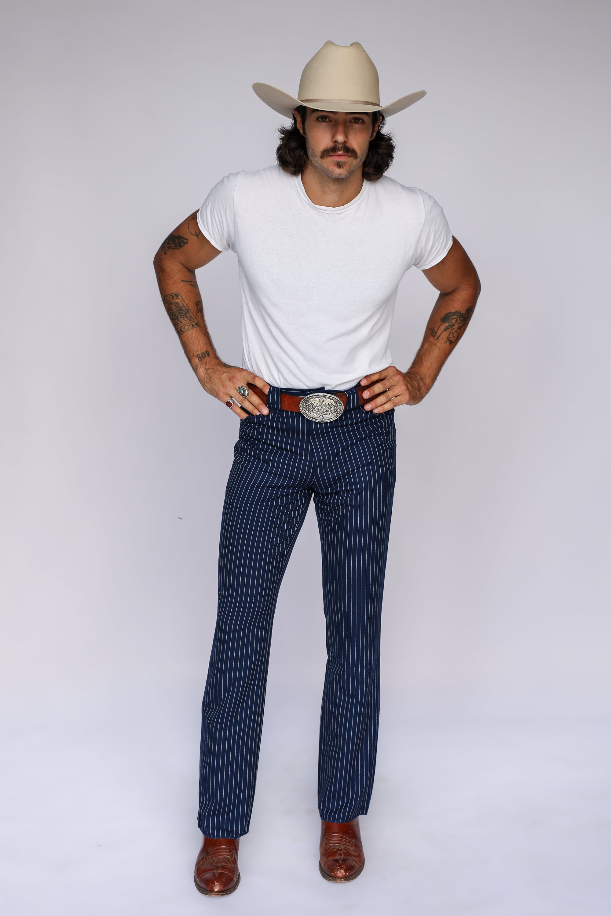 A man modeling a pair of dark navy blue pinstriped trousers with a large silver buckle belt, styled with a white t-shirt, brown boots, and a beige cowboy hat against a white studio background.