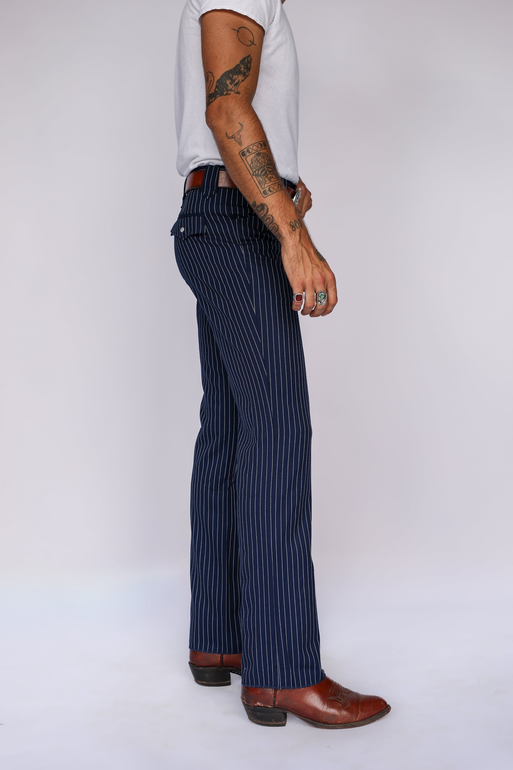 A side-profile view of a person wearing navy blue pinstriped trousers, a white t-shirt, and brown leather boots against a plain light gray studio background.