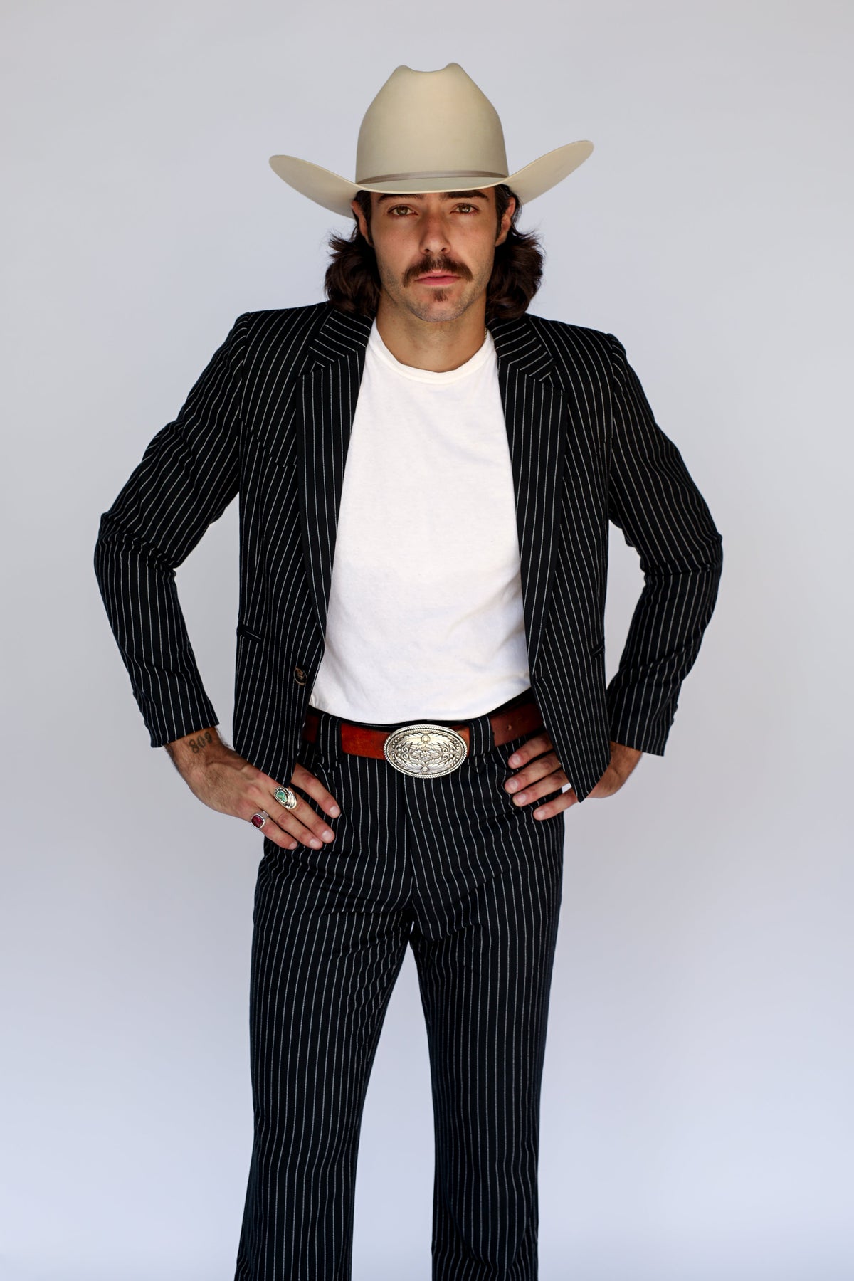 A man modeling a black and white pinstripe suit with a white t-shirt, a red belt with a large silver buckle, and a beige cowboy hat, posed against a light gray studio background.