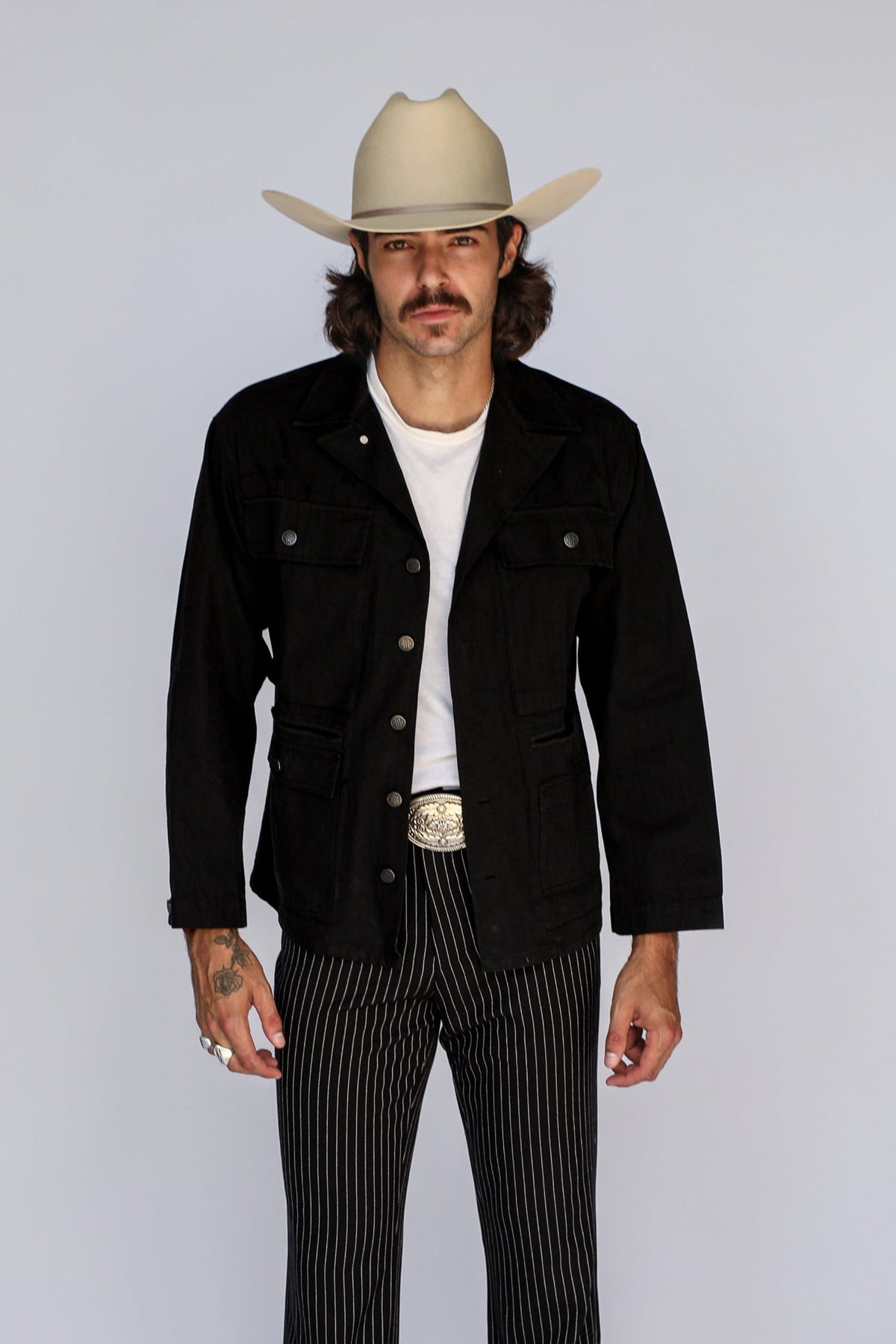 A man modeling a western-style outfit consisting of a black denim jacket, black and white pinstripe pants, a white t-shirt, a silver belt, brown leather shoes, and a beige cowboy hat, photographed against a light gray studio background.
