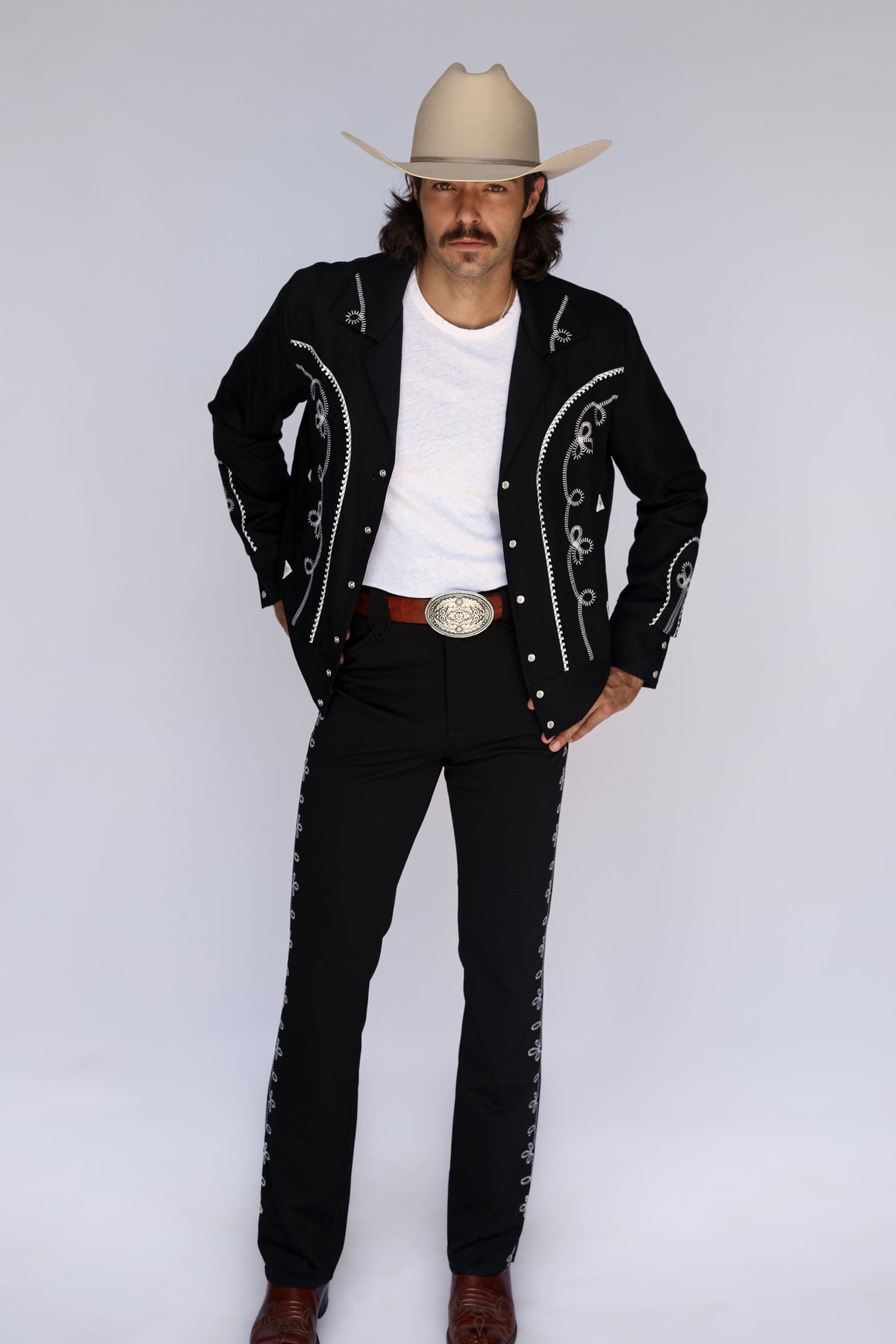 A man modeling a western-style outfit consisting of a black embroidered jacket, black pants, a white t-shirt, a brown leather belt with a large silver buckle, and a beige cowboy hat, photographed against a light gray studio background.
