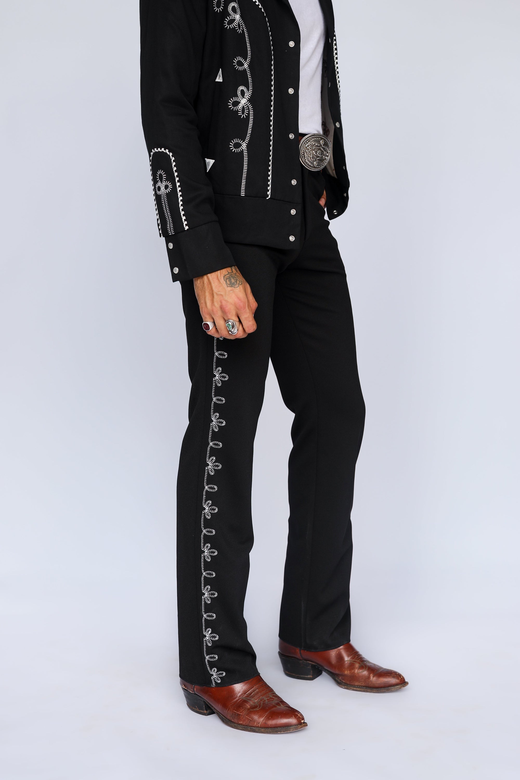 A person wearing a black western-style outfit with silver embroidered details, a white shirt, a large silver belt buckle, and brown leather cowboy boots, photographed against a light gray studio background.