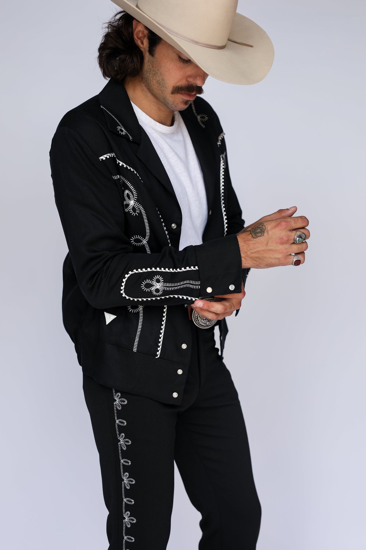 A man wearing a black western-style jacket and matching pants with white decorative embroidery, a white t-shirt, and a beige cowboy hat, photographed against a light gray studio background.