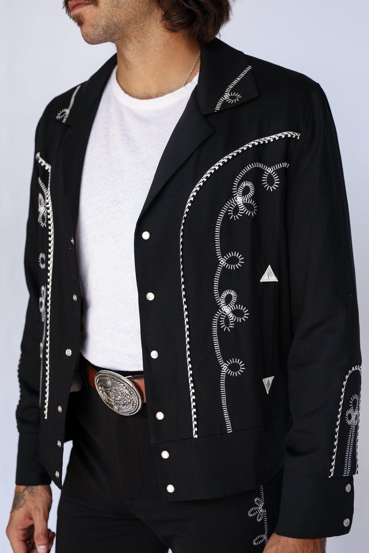 A man wearing a black western-style jacket with white embroidered details, a white t-shirt, and black pants with a large silver belt buckle, shown from the chin down against a light gray background.