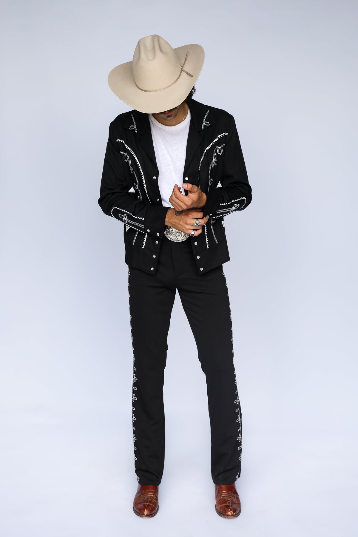 A man wearing a black embroidered western-style jacket and matching black pants with decorative stitching down the sides, a white t-shirt, brown boots, and a beige cowboy hat, standing against a light gray studio background.