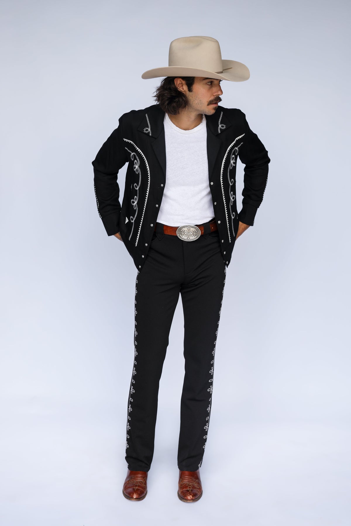 A man modeling a black charro-style jacket and matching pants with silver embroidery, a white t-shirt, a brown belt, a beige cowboy hat, and brown boots against a light gray studio background.