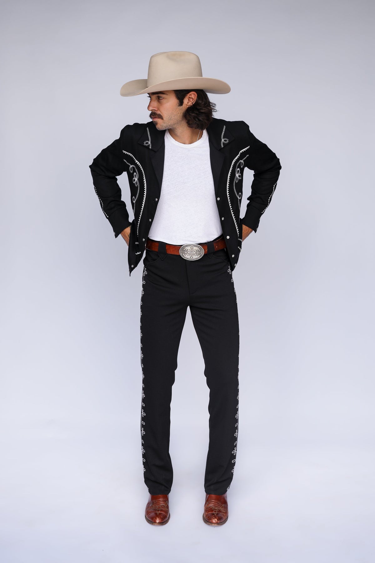 A man modeling a black western-style jacket and matching pants with silver embroidery, a white t-shirt, a brown belt, a beige cowboy hat, and brown boots, standing against a light gray studio background.