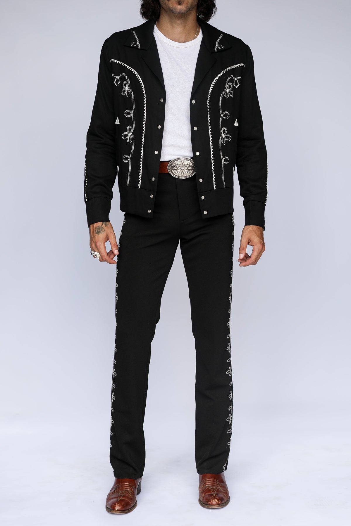 A man modeling a black western-style jacket and matching pants set with silver embroidered details, a white t-shirt, a silver belt, and brown leather shoes, against a light gray studio background.