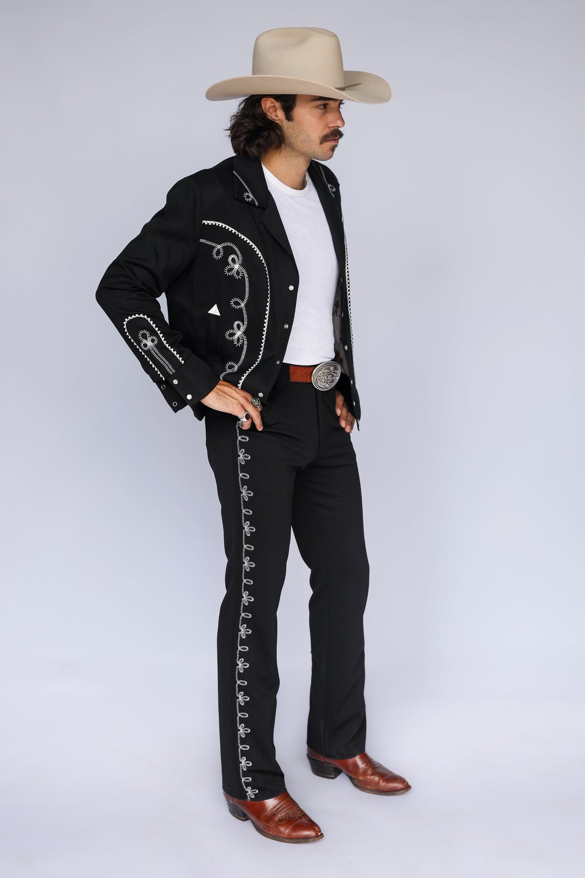 A man modeling a black charro-style jacket and matching pants with white embroidered trim, a white t-shirt, brown boots, and a beige cowboy hat against a light gray studio background.