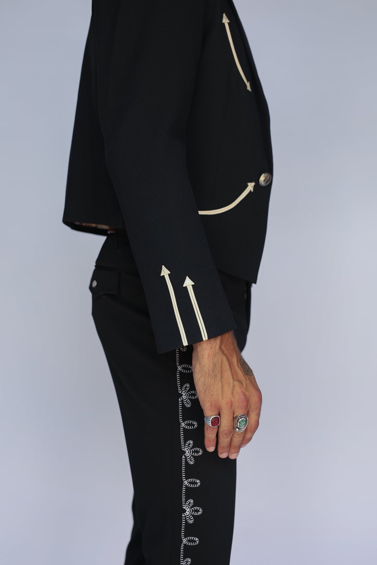 A close-up side view of a person wearing a black cropped blazer and matching black trousers, both detailed with contrasting beige embroidered arrow motifs and decorative trim. The person is also wearing several rings, and the background is a clean, light gray studio backdrop.