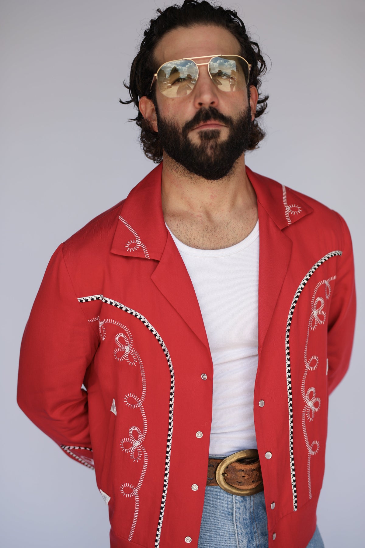 A man wearing a bright red western-style jacket with white embroidered scrollwork, a white t-shirt, blue jeans, and gold aviator sunglasses, photographed against a plain light gray background.