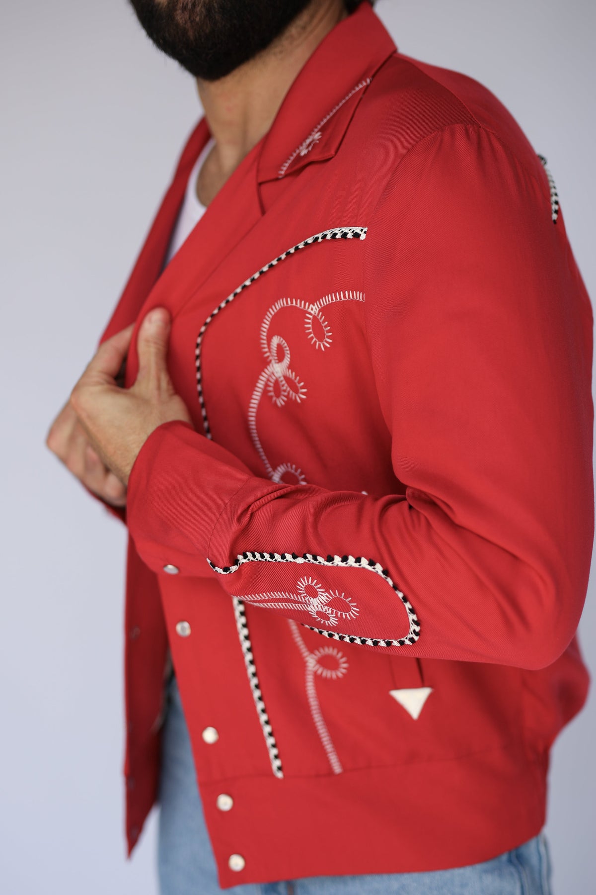 A close-up side view of a man wearing a bright red western-style jacket with white embroidered floral details and black-and-white checkered trim, paired with light blue jeans against a plain light gray background.